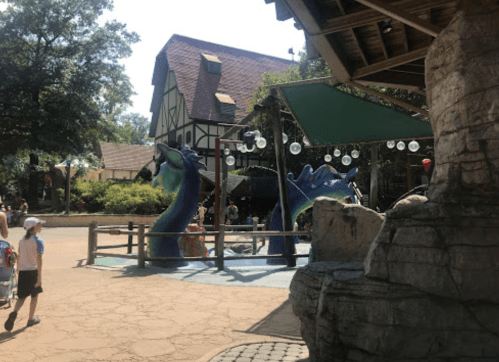 A whimsical water play area featuring blue dragon sculptures, surrounded by trees and a rustic building in the background.