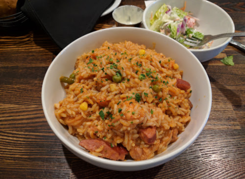 A bowl of flavorful rice with shrimp, sausage, and vegetables, garnished with herbs, alongside a small salad.