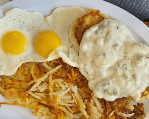 A plate of breakfast featuring two sunny-side-up eggs, hash browns, a biscuit, and sausage gravy.