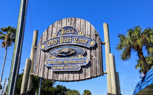 Sign for "Perk's Old Port Cove Seafood Restaurant" with a crab illustration, surrounded by palm trees and blue sky.