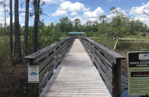 A wooden boardwalk leads through a natural area, surrounded by trees and water, with signs about fishing regulations.