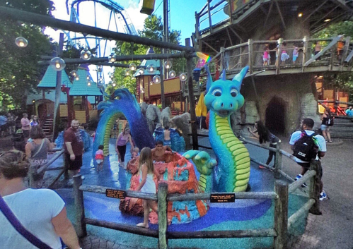 Colorful dragon fountain in a children's play area, with kids playing and adults nearby in a theme park setting.