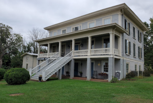 A large, two-story gray house with a porch, white stairs, and green lawn, surrounded by trees and shrubs.