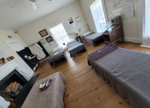 A simple room with wooden floors, featuring several beds, a fireplace, and framed artwork on the walls.