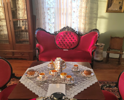 A vintage parlor with a red velvet sofa, ornate table, and a tea set arranged on a lace tablecloth.
