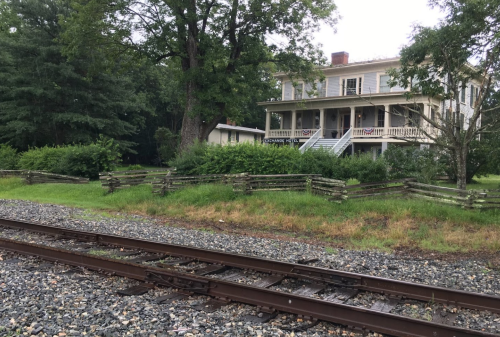 A historic house with a porch, surrounded by greenery, near a gravel railway track on a rainy day.