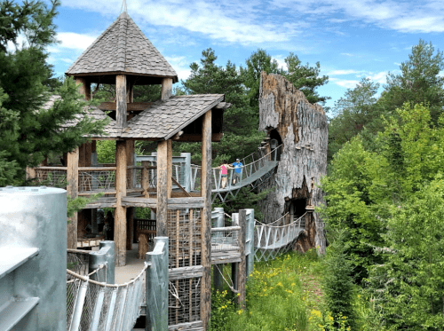 A wooden play structure with a tower and rope bridge, surrounded by trees and greenery under a blue sky.