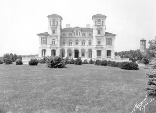 A large, historic mansion with towers, surrounded by greenery and a clear sky, captured in black and white.