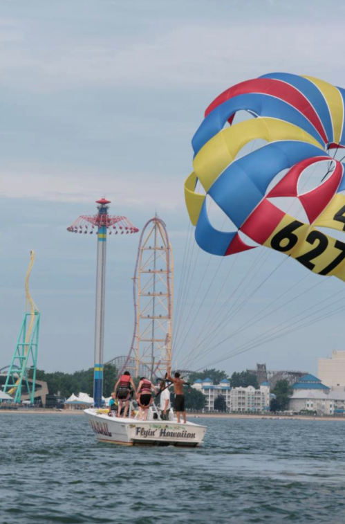 A boat with people parasailing, colorful parachute above, amusement park rides in the background, on a calm water surface.