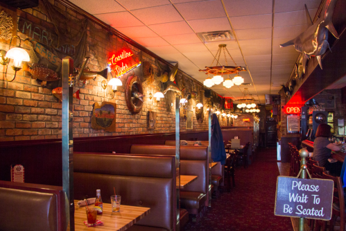 Cozy restaurant interior with brick walls, wooden booths, and vintage decor, featuring a "Please Wait to Be Seated" sign.