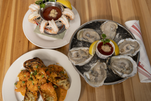 A plate of oysters with lemon and cocktail sauce, shrimp cocktail, and a dish of shrimp pasta on a wooden table.