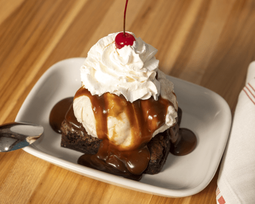A warm brownie topped with vanilla ice cream, caramel sauce, whipped cream, and a cherry on a white plate.