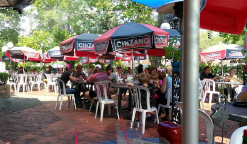 Outdoor dining area with people seated under colorful umbrellas at a restaurant, surrounded by greenery.