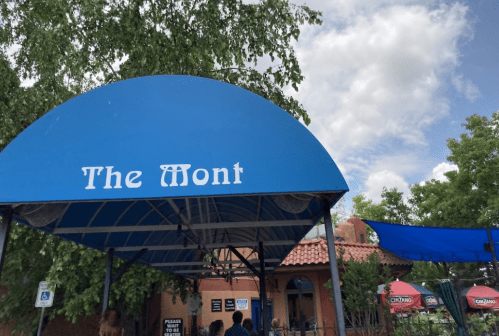 A blue awning with "The Mont" written on it, outside a restaurant with trees and a cloudy sky in the background.
