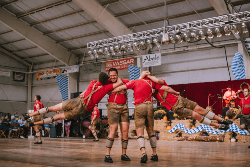Men in traditional attire perform a lively dance, lifting each other in a festive indoor setting.