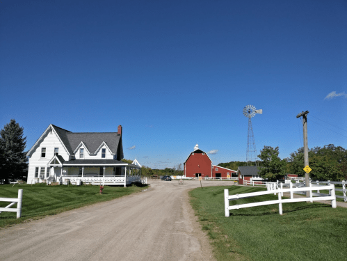 A scenic rural landscape featuring a white farmhouse, red barn, windmill, and clear blue sky.