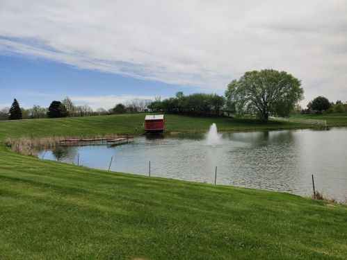 A serene pond with a red barn, fountain, and lush green grass under a blue sky with scattered clouds.