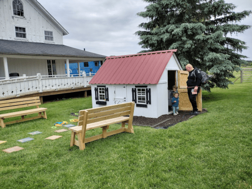 A child stands by a small playhouse while an adult approaches, with benches and greenery in the background.