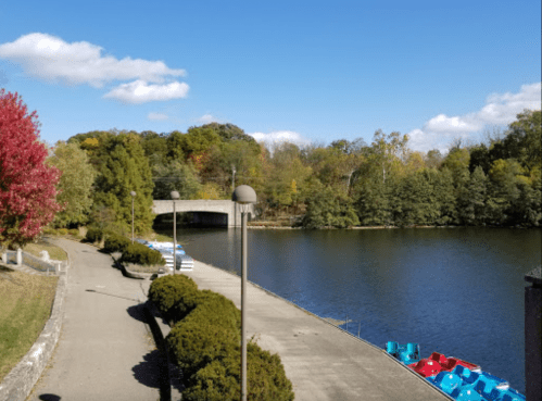 A serene lakeside view with colorful trees, a bridge, and paddle boats lined up along the shore under a clear blue sky.