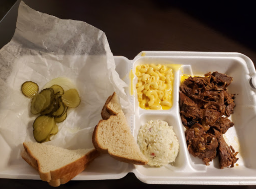 A takeout container with pulled beef, macaroni and cheese, coleslaw, pickles, and slices of bread.