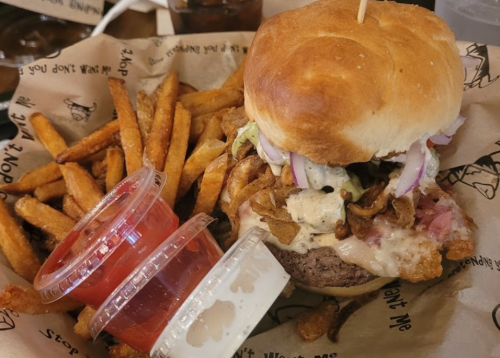 A juicy burger topped with sauce and onions, served with crispy fries and two dipping sauces in plastic cups.