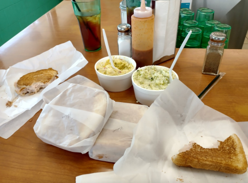 A table with two sandwiches wrapped in paper, two cups of coleslaw, and condiments, alongside a drink.