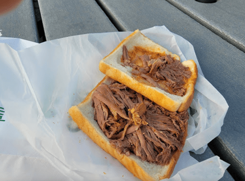 A sandwich with shredded beef on white bread, resting on a paper wrapper on a wooden table.