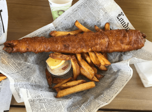 A large fried fish fillet served with golden fries and a lemon wedge on a bed of newspaper.