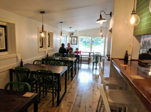 A cozy café interior with wooden tables, green chairs, and a large window letting in natural light.