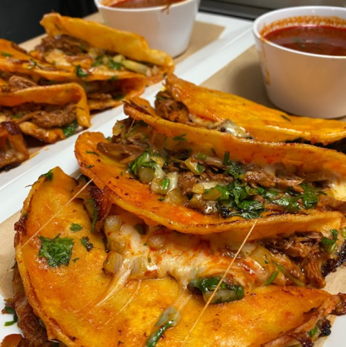 A close-up of stacked tacos filled with meat, cheese, and herbs, served with two small bowls of sauce.