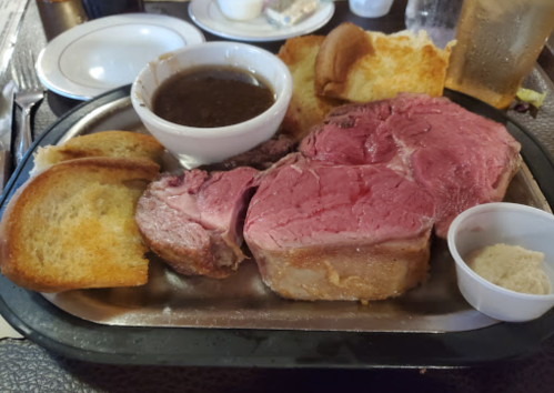 A plate with a large portion of pink prime rib, gravy, toast, and a small cup of horseradish on a metal tray.