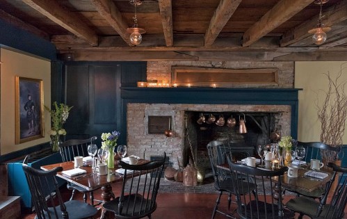 Cozy dining area with wooden beams, a fireplace, and tables set with glassware and flowers. Warm, inviting atmosphere.