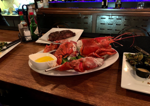 A platter of lobster with melted butter, accompanied by a steak and side dishes on a wooden bar table.