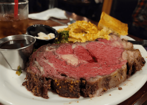 A plate featuring a large slice of prime rib, sides of greens, mashed potatoes, and a piece of toasted bread.