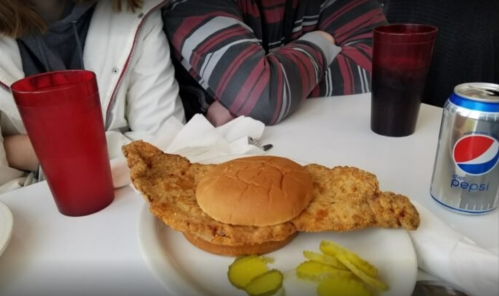 A large fried fish fillet sandwich on a plate, accompanied by pickles and a can of Pepsi, with people in the background.