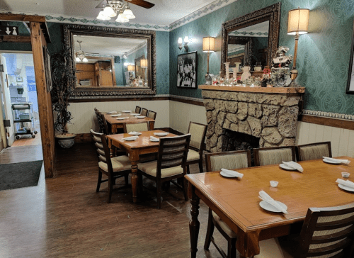 Cozy restaurant interior with wooden tables, chairs, and a stone fireplace, featuring green wallpaper and decorative mirrors.