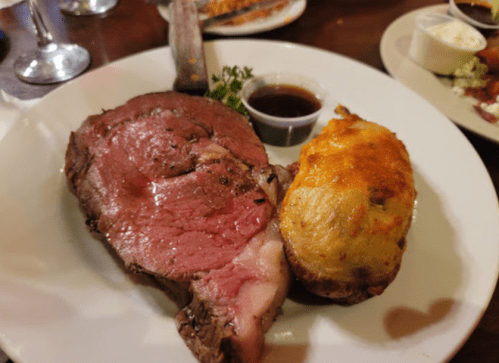 A plate featuring a juicy prime rib with a side of cheesy stuffed potato and a small cup of sauce.