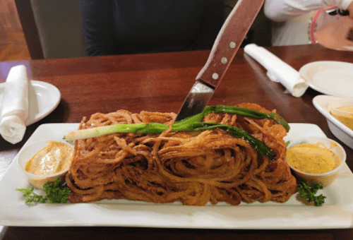 A large serving of crispy onion rings topped with a knife and garnished with green onions, accompanied by two dipping sauces.