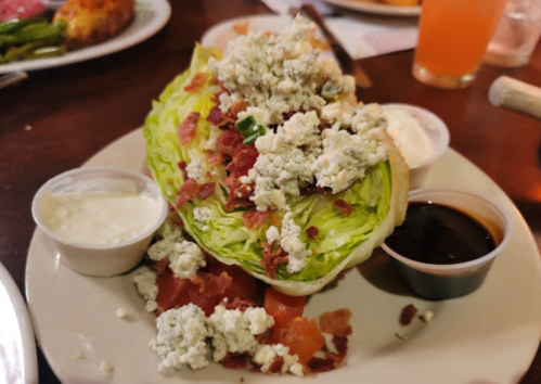 A wedge of lettuce topped with blue cheese, bacon, and diced tomatoes, served with ranch and balsamic dressings.