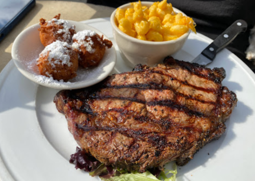 A grilled steak on a plate with macaroni and cheese and three fried corn fritters dusted with powdered sugar.
