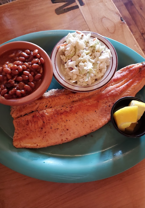A plate with two grilled fish fillets, coleslaw, baked beans, and a lemon wedge on a turquoise platter.