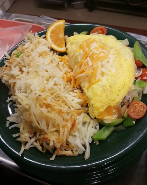 A green plate with a cheese omelet, hash browns, and a side of sautéed vegetables and cherry tomatoes, garnished with an orange slice.