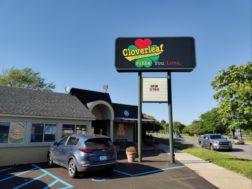 Exterior of Cloverleaf Pizza restaurant with a sign reading "Open 12 to 8" and a parked car in front.