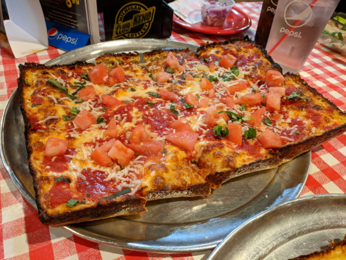 A rectangular pizza topped with cheese, diced tomatoes, and herbs on a metal tray, set on a red and white checkered tablecloth.