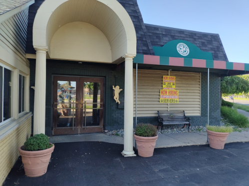 Exterior of a building with a sign reading "Now Hiring Experienced Servers" and potted plants at the entrance.