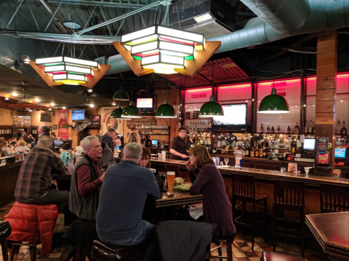 A lively bar scene with patrons at tables, a long bar, and colorful pendant lights overhead.
