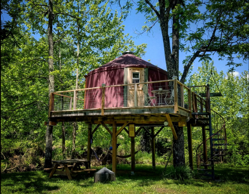 A cozy yurt on stilts surrounded by lush green trees, featuring a wooden deck and spiral staircase.