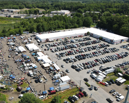 Aerial view of a busy outdoor market with numerous cars parked and vendor tents set up in a large lot surrounded by greenery.