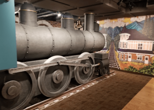 A vintage steam locomotive model displayed indoors, with a mural of mountains and a train track in the background.
