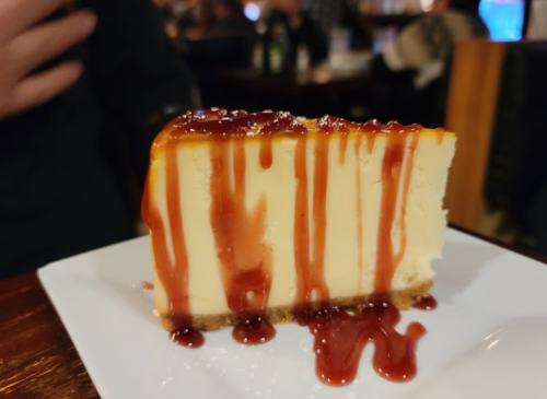 A slice of cheesecake drizzled with caramel sauce on a white plate, set in a dimly lit restaurant.